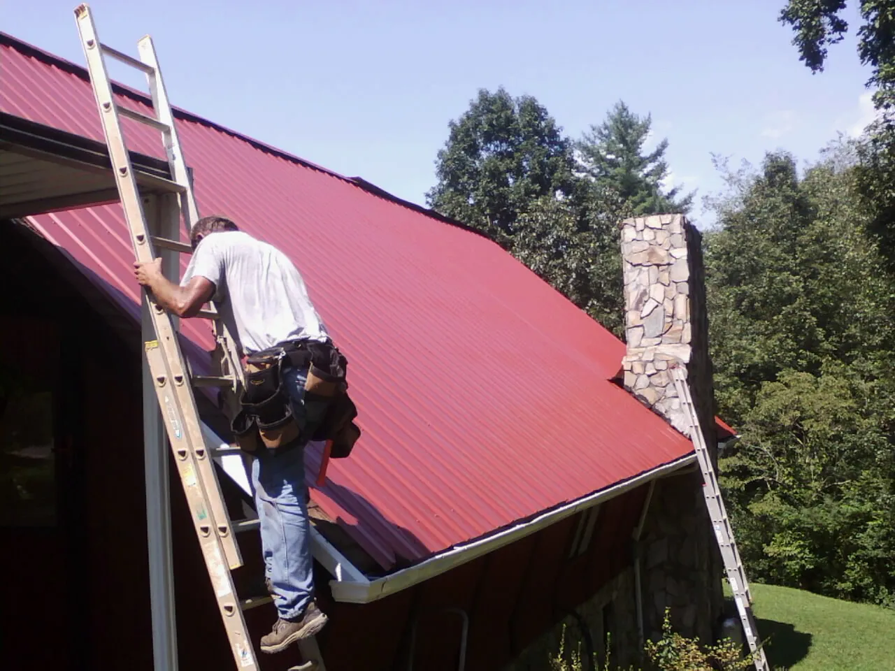 Roofer on a ladder beside a red metal roof and stone chimney — Soffit & Fascia Repair by Roofing Brothers in Velma